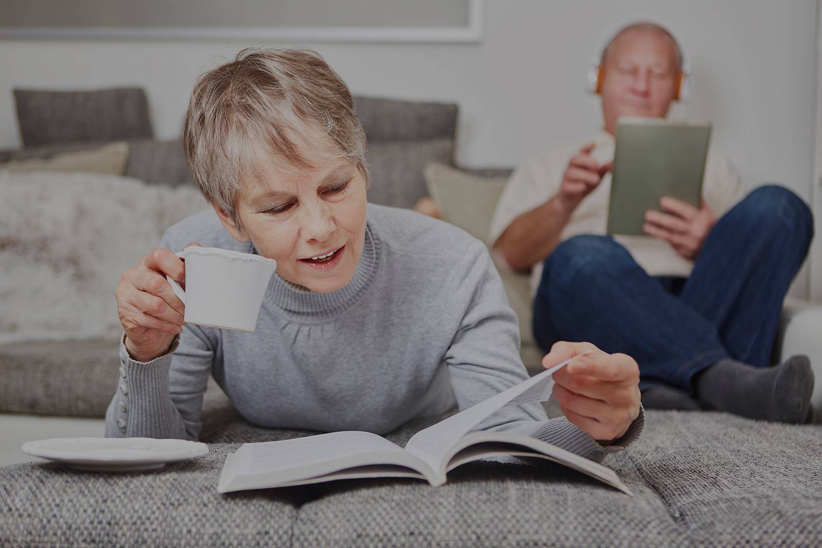 Fotografía de una mujer mayor leyendo mientras toma un café en el sofá de su casa con su pareja al fondo